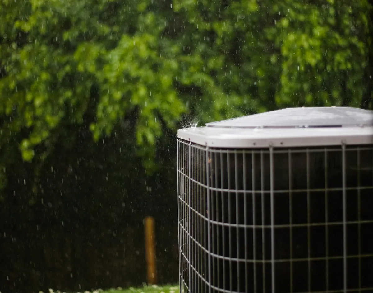 An outdoor air-conditioning unit in a backyard during a rain shower, with green trees behind it.