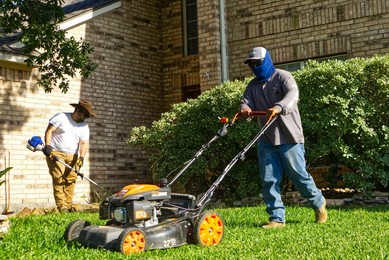 Two workers on a front lawn in front of a brick house: one pushes a mower, the other runs a string trimmer along the bed.